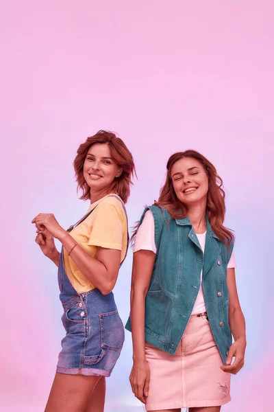 Two joyful attractive young girls, twin sisters smiling, dancing while posing together isolated over pink background — Stock Photo, Image