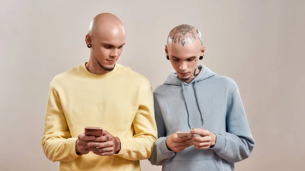 Zwei junge kaukasische Zwillingsbrüder in Freizeitkleidung mit Tattoos und Piercings auf Smartphones, während sie isoliert vor beigem Hintergrund stehen — Stockfoto