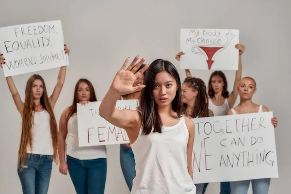 Beyaz tişörtlü genç Asyalı kadın kameraya bakıyor, gösteriyor, dur hareketi yapıyor. Arka planda protesto pankartları taşıyan bir grup kadın — Stok fotoğraf