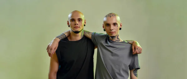 Verdadeiros amigos. Imagem horizontal de dois jovens irmãos gêmeos com tatuagens e piercings abraçando e olhando para a câmera enquanto posam juntos em estúdio, bandeira panorâmica — Fotografia de Stock