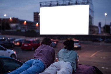 Burada iyi vakit geçirin. İki kız bir arabanın çatısında uzanıyor, akşam büyük beyaz bir ekranla açık hava sinemasında film izlerken eğleniyorlar.