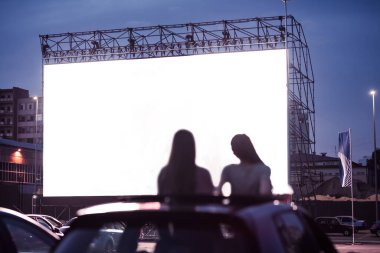 Açık hava sinemasında büyük beyaz ekranlı bir film izlerken arabada oturan iki kadın arkadaşın arka görüntüsü.