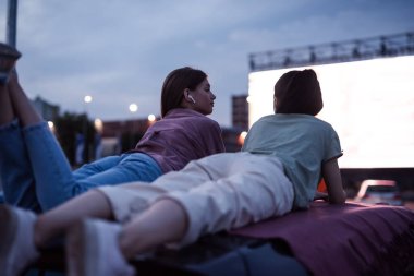İki kız bir arabanın çatısında uzanıp büyük beyaz ekranlı açık hava sinemasında film izlerken eğleniyorlar.
