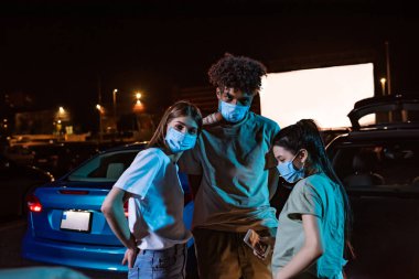Koruyucu maskeler takan üç farklı arkadaş büyük bir ekranın önüne park etmiş arabanın yanında duruyorlar. Açık hava sinemasında film izleyen gençler.