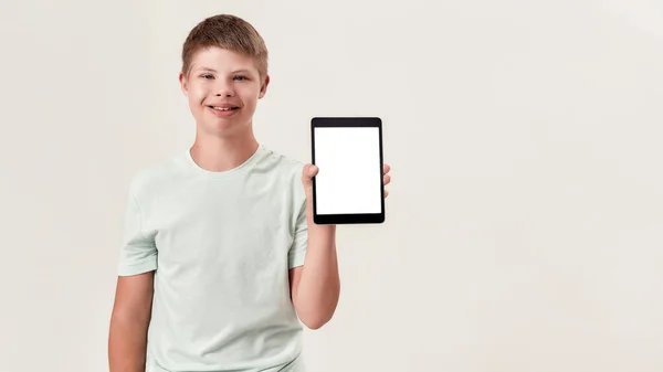 Glücklicher behinderter Junge mit Down-Syndrom lächelt, hält und zeigt Tablet-PC mit leerem Bildschirm, während er isoliert vor weißem Hintergrund steht — Stockfoto