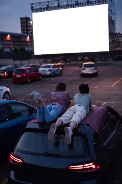 Rückansicht von zwei Freunden, die auf dem Dach eines Autos liegen, während sie sich einen Film in einem Open-Air-Kino ansehen. Autos parken vor einer großen weißen Leinwand, um Filme anzusehen — Stockfoto