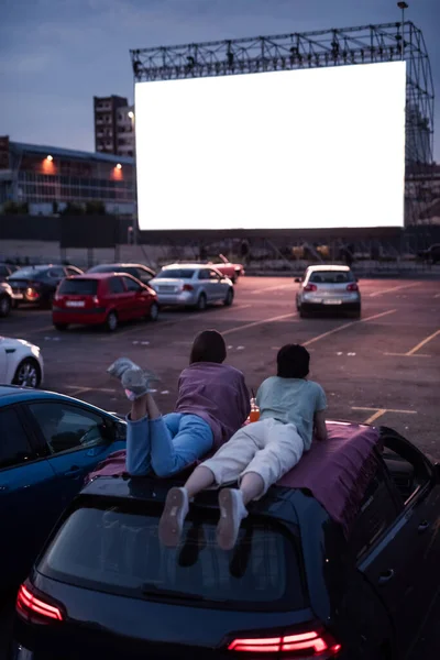映画をお楽しみください。オープンエアの映画館で映画を見ながら、車の屋根の上に横たわっている2人の友人のリアビュー。映画を見るために大きな白い画面の前に駐車車 — ストック写真