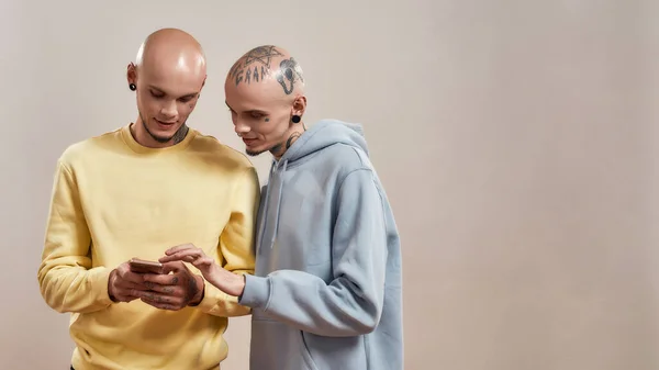 Jag tittar på video. Två unga kaukasiska tvillingbröder i casual wear med tatueringar och piercingar med hjälp av smartphone samtidigt stå isolerade över beige bakgrund — Stockfoto