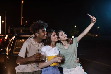 Neşeli çeşitli genç arkadaşlar birlikte selfie çekerken gülümsüyorlar, büyük bir ekranın önünde duruyorlar, açık hava sinemasında film izlemeye hazırlar.