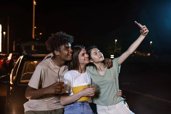 Alegres y diversos jóvenes amigos sonriendo mientras se toman una selfie juntos, de pie frente a una pantalla grande, listos para ver una película en un cine al aire libre — Foto de Stock