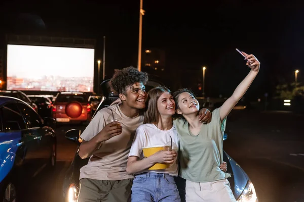 Joyful diverso jóvenes mejores amigos posando mientras se toman una selfie juntos, de pie frente a una pantalla grande, listo para ver una película en un cine al aire libre —  Fotos de Stock
