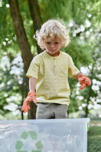 Cute little caucasian boy wearing rubber gloves collecting plastic waste in the forest or park, plastic bottles in recycle Stock Photo