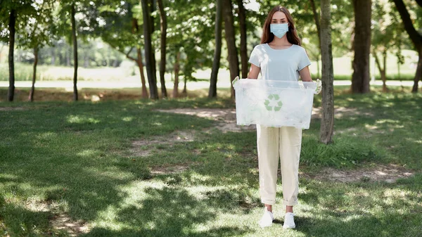 Mujer joven, activista ecológica que usa mascarilla protectora que sostiene la papelera de reciclaje, mirando a la cámara mientras recoge desechos plásticos en el bosque — Foto de Stock