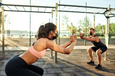 Koruyucu maskeli genç sporcular spor sahasında çömelme yapıyorlar.