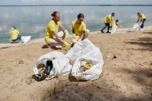 Dos bolsas de basura en la arena, grupo de jóvenes eco activistas o voluntarios en uniforme recogiendo residuos plásticos y basura en la playa — Foto de Stock