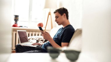 Genç adam evde kanepede otururken dizüstü bilgisayar ve cep telefonu kullanıyor. Teknoloji, akıllı telefon bağımlılığı konsepti