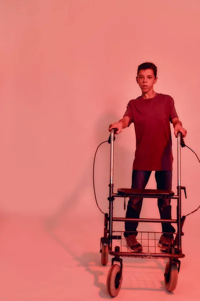 Full length shot of teenaged disabled boy with cerebral palsy looking at camera, taking steps with his walker isolated over red light background — Stock Photo, Image