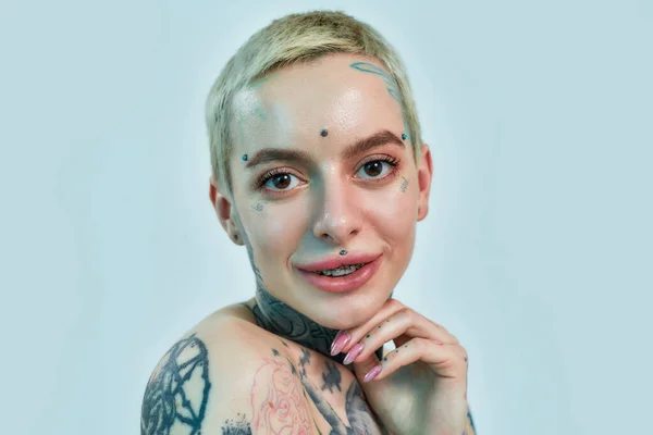 Κοντινό πλάνο μιας λευκής γυναίκας με τατουάζ με σκουλαρίκια και αγκύλες που κοιτάζει σε μια κάμερα σε μπλε φόντο — Φωτογραφία Αρχείου