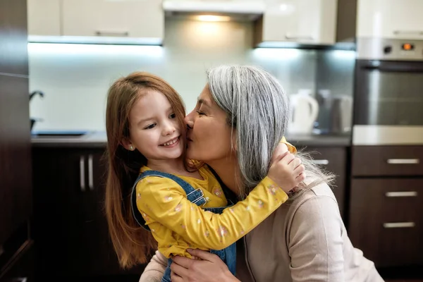 Baci. Ritratto di nonna felice che abbraccia e bacia la sua graziosa nipotina prescolare, in piedi insieme a casa — Foto Stock