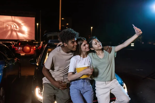 Alegres y diversos jóvenes amigos sonriendo mientras se toman una selfie juntos, de pie frente a una pantalla grande, listos para ver una película en un cine al aire libre —  Fotos de Stock