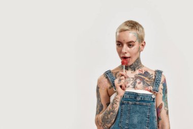 Dövme ve piercing. Üzerinde kot pantolon olan, kameraya bakarken lolipop yalayan, piercing yaptırmış, dövmeli bir kadın.