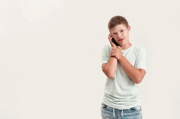 Portrait of teenaged disabled boy with Down syndrome looking at camera while talking on the phone, standing isolated over white background — Stock Photo, Image
