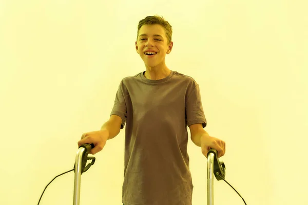 Cheerful teenaged disabled boy with cerebral palsy smiling at camera, taking steps with his walker isolated over yellow light background — Stock Photo, Image