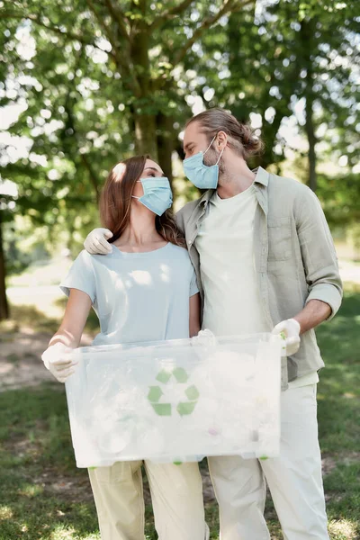 Junges Paar mit Mundschutz hält Papierkorb in der Hand, umarmt sich und schaut sich an, sammelt gemeinsam Plastikmüll im Wald wegen COVID19 — Stockfoto