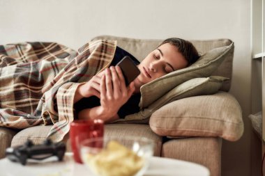 Genç adam dinlenirken cep telefonunu kullanıyor, evdeki kanepede yatıyor. Akıllı telefon bağımlılığı, ilgisizlik ve hüsran kavramı.