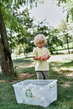 Ormanı temizlerken veya parkı temizlerken elinde plastik şişe tutan beyaz bir çocuğun dikey görüntüsü. Çöp kutusunun yanında duruyor.