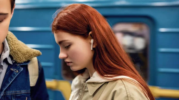 Retrato de cerca de una adolescente con auriculares inalámbricos mirando hacia abajo, escuchando música mientras está parada en la estación de metro —  Fotos de Stock