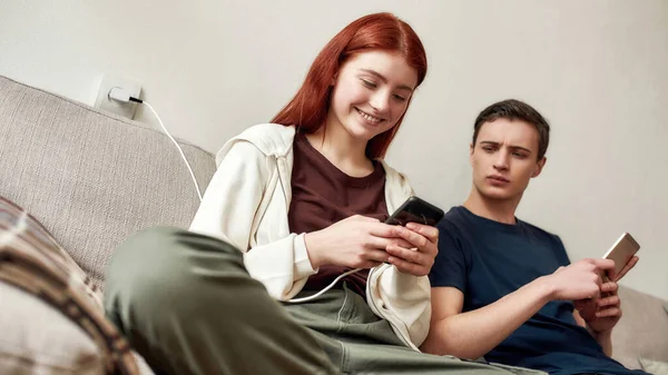Un par de adolescentes sentados en el sofá de casa, usando el teléfono móvil. Un tipo mira celosamente la pantalla del teléfono de su novia. Sin confianza, adicción a los teléfonos inteligentes, concepto de problema de comunicación —  Fotos de Stock