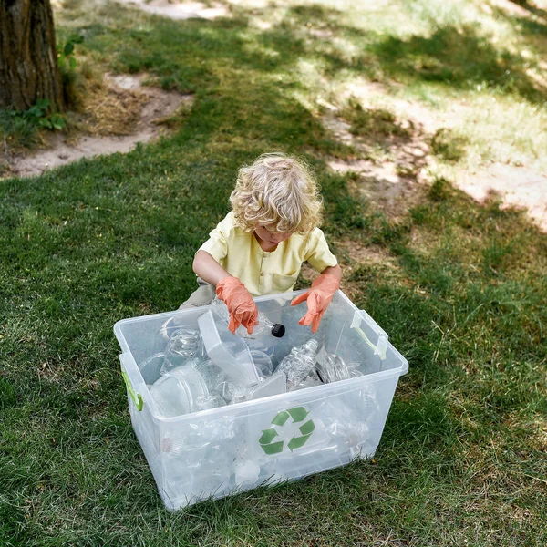 Lindo niño caucásico con guantes de goma arrojando residuos de plástico en la papelera de reciclaje mientras limpia en el bosque o el parque — Foto de Stock