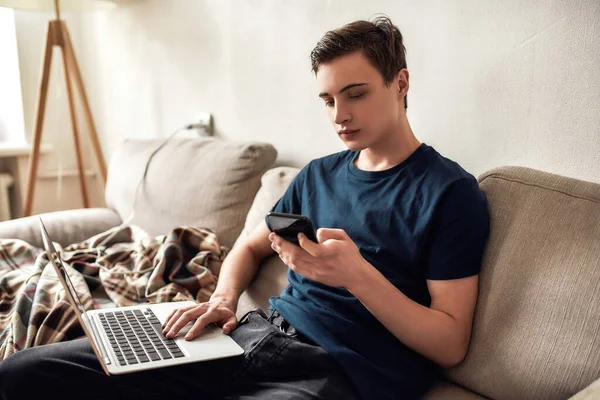Chico joven que usa el ordenador portátil y el teléfono móvil mientras pasa tiempo sentado en el sofá en casa. Tecnología, concepto de adicción a teléfonos inteligentes —  Fotos de Stock