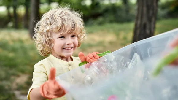 Salva nuestro planeta. Lindo niño feliz sosteniendo papelera de reciclaje con residuos de plástico y sonriendo mientras limpia de basura en el bosque o parque — Foto de Stock