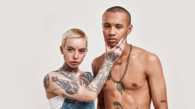 Çok kültürlü bir ilişki. Dövmeli, beyaz, piercingli bir kadın koyu tenli, üstsüz bir adama dokunuyor. İkisi de kameraya bakıyor.