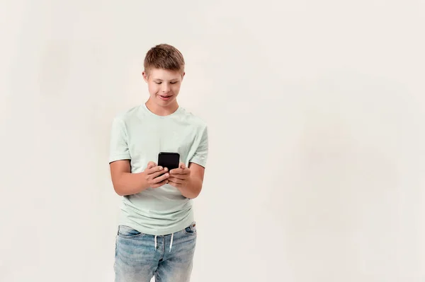 Niño discapacitado adolescente con síndrome de Down sosteniendo el teléfono inteligente, mirando a la pantalla mientras está de pie aislado sobre fondo blanco — Foto de Stock