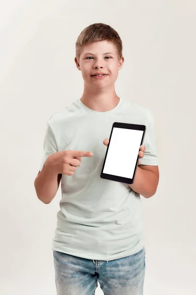 Niño discapacitado feliz con síndrome de Down sonriendo, sosteniendo y apuntando a la tableta PC con pantalla en blanco mientras está de pie aislado sobre fondo blanco —  Fotos de Stock