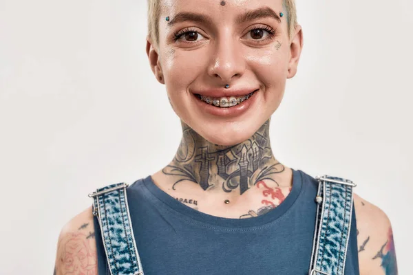 Retrato de uma mulher branca com tatuagens e piercing olhando para uma câmera sorrindo usando suportes e uma ganga geral — Fotografia de Stock