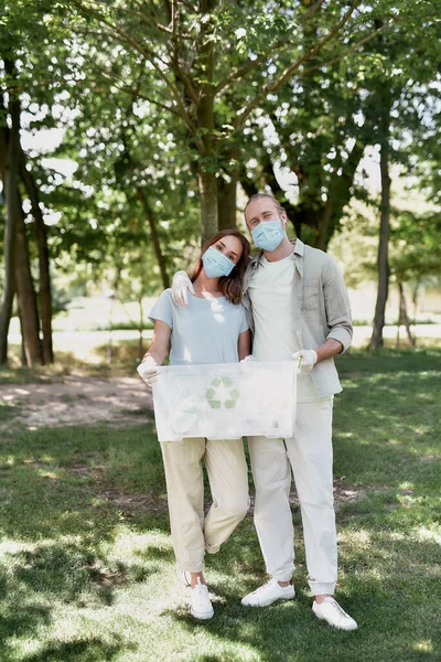 Pareja joven con mascarillas protectoras con papelera de reciclaje abrazando y mirando a la cámara, recogiendo residuos plásticos juntos en el bosque debido a la pandemia de COVID19 — Foto de Stock