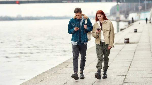 Colpo completo di due adolescenti totalmente assorbiti nei loro smartphone, ignorandosi a vicenda mentre camminano lungo il lungofiume insieme — Foto Stock
