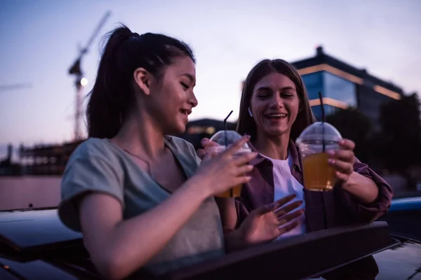 Dos chicas que se ven alegres, sosteniendo bebidas mientras están sentadas en el coche y viendo una película en un cine al aire libre por la noche — Foto de Stock