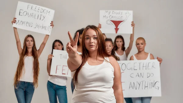 Beyaz tişörtlü beyaz bir kadın kameraya bakarak barış işareti gösteriyor. Arka planda protesto pankartları taşıyan bir grup kadın — Stok fotoğraf
