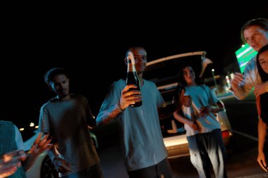 Genç, dövmeli bir adam kameraya bakıp gülümseyen ve elinde bir şişe şampanya tutan çok kültürlü arkadaşları için açılmış.