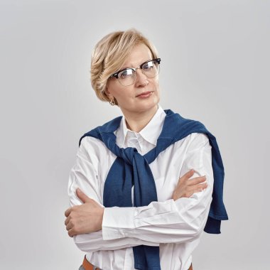 İş elbisesi ve gözlük takmış zarif orta yaşlı beyaz kadın portresi poz verirken kameraya bakıyor, gri arka planda kolları çaprazlanmış duruyor.