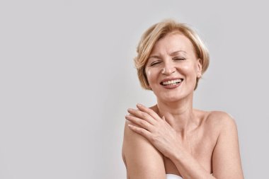 Güzel orta yaşlı bir kadının portresi kameraya gülüyor, omzuna dokunuyor, gri arka planda izole bir poz veriyor. Güzellik, cilt bakımı