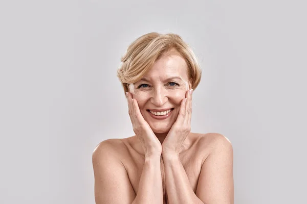 Retrato de una hermosa mujer de mediana edad desnuda mirando emocionada, sonriendo a la cámara, tocándose las mejillas mientras posaba aislada sobre un fondo gris. Belleza, concepto de cuidado de la piel —  Fotos de Stock