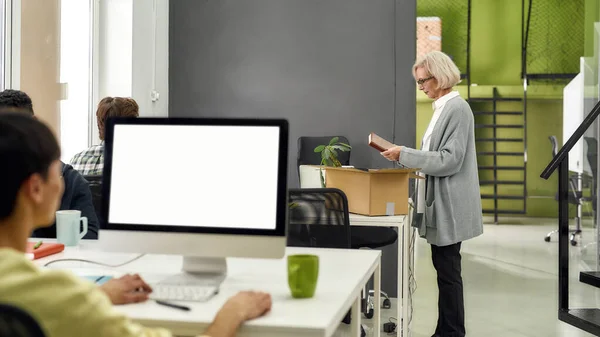 New employee aged woman, senior female intern holding her notebook while pulling personal belongings out of open box, preparing for first day at work — Stock Photo, Image