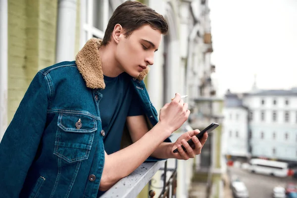 Πορτρέτο του έφηβου καπνίσματος και γραπτών μηνυμάτων χρησιμοποιώντας το κινητό του τηλέφωνο, ενώ στέκεται στο μπαλκόνι — Φωτογραφία Αρχείου