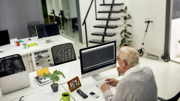 Back view of aged man, senior intern in wireless earphones looking focused while typing, using pc, sitting at desk, working in modern office — Stock Photo, Image